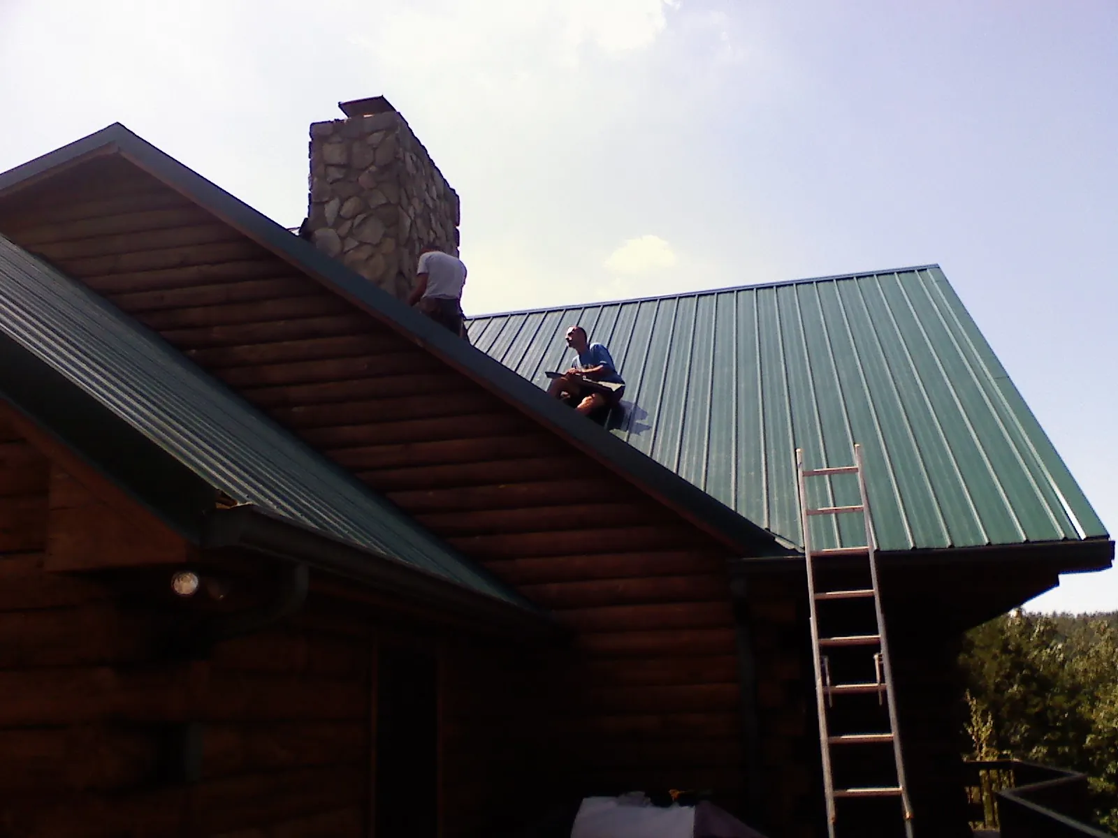 Roofers installing green metal panels beside a stone chimney on a log home in Tremont